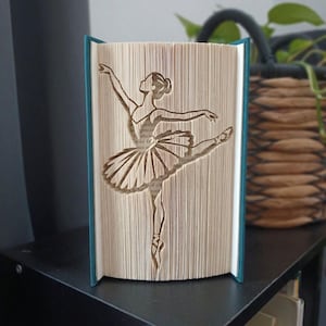 Può includere: Una scultura in libro piegato che presenta la sagoma di una ballerina in posa di danza. Le pagine sono piegate con precisione per creare l'immagine di una ballerina con le braccia tese e una gamba sollevata, su una copertina color ottanio.