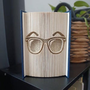 May include: A folded book art sculpture featuring a pair of glasses with eyebrows created by precisely folding the individual pages of a book. The book has a dark blue cover and the pages are cream-colored with visible black printed text.