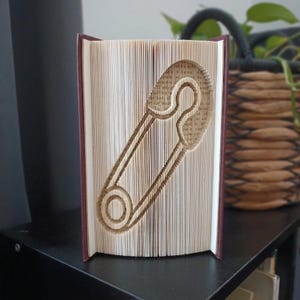 Op de afbeelding: Een gevouwen boekkunstsculptuur met een veiligheidsspeldontwerp, gemaakt door de individuele pagina's van een hardcover boek nauwkeurig te vouwen. De pagina's zijn crèmekleurig met zichtbare zwarte tekst en de kaft is donkerrood.