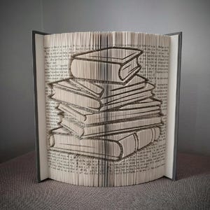 May include: A unique book art piece featuring a stack of books carved into the pages of an open book. The pages are folded to create the image of a stack of books. The book has a gray cover and is displayed on a gray surface.