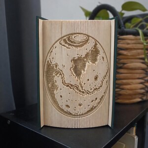 Puede incluir: Una escultura de libro con páginas plegadas que forman un relieve circular detallado de la superficie lunar. Las páginas son de color crema con bordes oscuros, y el diseño muestra cráteres y un terreno lunar texturizado.