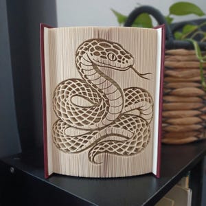 May include: A book sculpture featuring a snake design created by folding the pages of a hardback book. The pages are precisely manipulated to form the detailed silhouette of a coiled snake with a visible tongue, showcasing the paper edges.