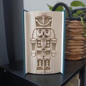 Op de afbeelding: Een gevouwen boekkunstsculptuur met een notenkrakerontwerp, gemaakt door de pagina's van een boek nauwkeurig te vouwen. De sculptuur toont de figuur van een soldaat met een hoge hoed, een snor en een uniform, tegen een donkerblauwe kaft.