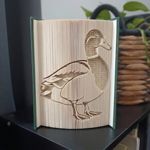 Peut inclure: Une sculpture en livre plié représentant la silhouette d'un canard, réalisée en pliant précisément les pages individuelles d'un livre à couverture rigide. Les pages sont couleur crème avec du texte noir et la couverture est vert foncé.