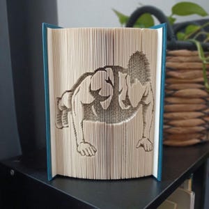 May include: A book sculpture featuring folded pages that create a three-dimensional silhouette of a person performing a push-up. The book has a teal cover and the interior pages are cream-colored, showing the intricate folds forming the figure.