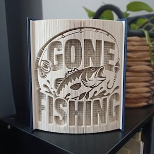 May include: A folded book art sculpture featuring the text GONE FISHING with a graphic of a fish and a fishing rod. The pages are precisely folded to create the three-dimensional lettering and imagery, showcasing the printed text of the book.
