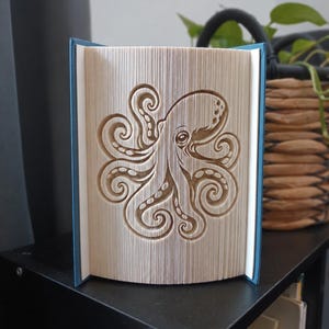 Può includere: Una scultura realizzata con la tecnica del libro piegato che presenta un dettagliato disegno di un polpo, creato piegando con precisione le singole pagine di un libro con copertina rigida. Il libro ha una copertina blu e le pagine formano un polpo.