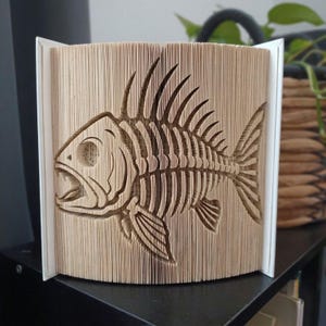 Può includere: Una scultura realizzata con un libro piegato che raffigura un dettagliato scheletro di pesce. Le pagine sono piegate con precisione per creare la forma tridimensionale della gabbia toracica, della testa e delle pinne, con il colore beige.