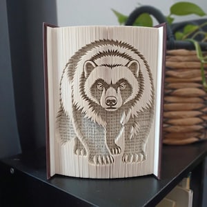Peut inclure: Une sculpture en livre plié représentant un ours détaillé, créée en pliant précisément les pages individuelles d'un livre à couverture rigide. L'ours est représenté de face avec une texture de fourrure et des traits faciaux visibles.