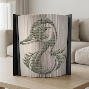 Peut inclure: Sculpture de livre décorative représentant un cygne. Le cygne est créé en pliant les pages d'un livre, l'image étant visible sur les bords. Le livre est maintenu dans un cadre noir et repose sur une surface en bois de couleur claire. Dimensions: environ 20 cm.