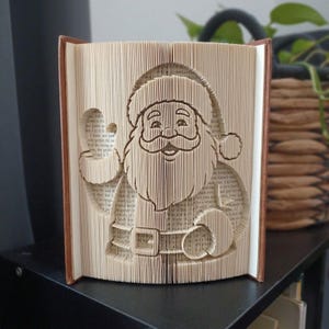 May include: A folded book art sculpture featuring a detailed silhouette of Santa Claus with a hat, beard, and belt. The pages of the book are precisely folded to create the three-dimensional image of the character, showing visible text on the paper.