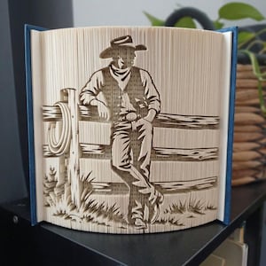 May include: A folded book art sculpture featuring a detailed silhouette of a cowboy wearing a hat and vest, leaning against a wooden fence with a coiled rope hanging on a post. The design is created by precisely folding the pages of a hardback book.