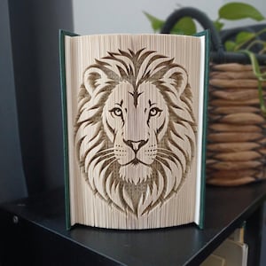 May include: A folded book art sculpture featuring a detailed lion face design created by precisely folding the individual pages of a hardback book. The book has dark green covers and the pages are cream-coloured with visible text throughout.