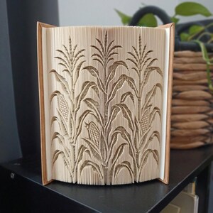 May include: A book sculpture featuring intricate folded pages that form a three-dimensional design of corn stalks with visible ears of corn. The pages are cream-colored and arranged to create a detailed relief pattern of the plants.