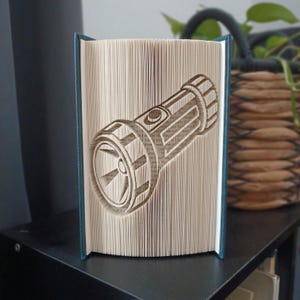 Può includere: Un libro con le pagine piegate per creare una scultura tridimensionale a forma di torcia. Il libro è posizionato in verticale e mostra l'intricato design in carta piegata della torcia sul bordo anteriore, con copertine blu scuro.