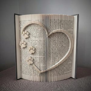 May include: A book with pages folded to create a heart shape and paw prints. The book is open, revealing the folded pages that form the heart and paw prints. The book has a gray cover and is sitting on a gray surface.