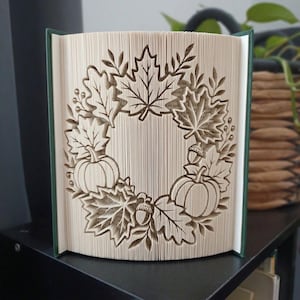 May include: A folded book art sculpture featuring a wreath design created from the pages of a book. The intricate paper folding displays a central circular pattern of maple leaves, two pumpkins, and acorns, showcasing the cream-colored paper edges.