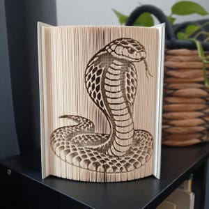 Può includere: Una scultura realizzata con un libro piegato che raffigura un cobra dettagliato. Le pagine del libro sono piegate con precisione per creare la forma tridimensionale di un cobra con il cappuccio aperto e la lingua fuori, evidenziando i bordi.