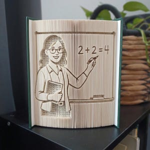 May include: A folded book art sculpture featuring a teacher figure with glasses holding a book, standing before a chalkboard with the equation 2+2=4 written on it. The sculpture is created from the pages of a book with a dark green cover.