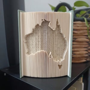 May include: A book sculpture featuring pages folded to create the distinct shape of the Australian continent. The book has a light green cover and the interior pages are cream-coloured with visible black printed text throughout the folded design.