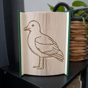 Op de afbeelding: Een boeksculptuur met een gevouwen paginadesign dat het silhouet van een meeuw vormt. Het boek heeft een donkergroene kaft en de pagina's zijn zo gerangschikt dat ze de gedetailleerde omtrek van de vogel tonen, inclusief de kop, vleugels en poten.