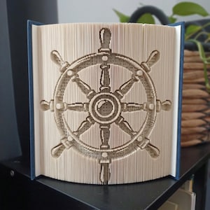 Op de afbeelding: Een gevouwen boekkunstsculptuur met een scheepsroerontwerp, gemaakt door de pagina's van een hardcover boek nauwkeurig te vouwen. De crèmekleurige papieren pagina's vormen de complexe cirkelvormige structuur van het nautische stuurwiel.