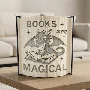 May include: A book sculpture with the words "BOOKS are MAGICAL" carved into the pages. A dragon illustration is centered on the book, with wings spread and sitting on an open book. The book has black edges and is displayed on a wooden surface.