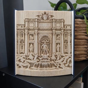 Peut inclure: Une sculpture de livre composée de pages pliées formant une représentation tridimensionnelle détaillée de la fontaine de Trevi, incluant des figures centrales, des colonnes architecturales et des sculptures, avec des couvertures bleu foncé.