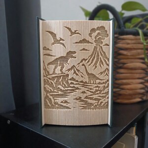May include: A folded book art sculpture featuring a prehistoric scene with a dinosaur standing on a cliff, a long-necked dinosaur near a river, two flying pterosaurs, and an erupting volcano with smoke clouds, all created from the pages of a book.