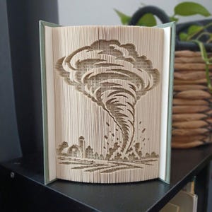 Può includere: Una scultura realizzata con un libro piegato che presenta un design dettagliato di un tornado creato piegando con precisione le pagine. La scultura raffigura un vortice che si estende sopra una piccola fattoria con un silo e una recinzione.