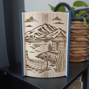 Op de afbeelding: Een gevouwen boekkunstsculptuur met een landschap met een bergketen, een kleine hut en een traditionele rieten boot op het water. De pagina's zijn nauwkeurig gevouwen om een driedimensionaal beeld te creëren met blauwe boekomslagen.