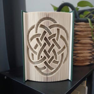 May include: A book sculpture featuring intricate folded pages that form a complex, symmetrical Celtic knot pattern. The book has dark green covers and the pages are cream-colored, creating a detailed three-dimensional design through precise paper folding techniques.