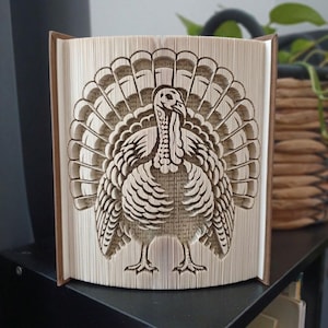 Peut inclure: Une sculpture de livre avec un design de pages pliées formant la silhouette d'une dinde avec une queue en éventail. Les pages sont de couleur crème avec du texte imprimé noir visible, et le livre est posé debout avec ses couvertures ouvertes.