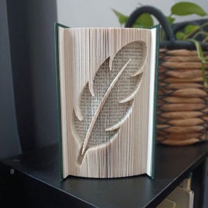 Puede incluir: Una escultura de libro con páginas dobladas que forman la silueta de una pluma. El libro tiene una cubierta de color verde oscuro y se mantiene en posición vertical, mostrando la técnica de plegado de papel del diseño central.