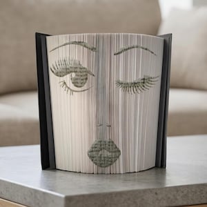 Puede incluir: Escultura decorativa de libro con diseño de rostro guiñando un ojo. El rostro se crea doblando las páginas de un libro, con detalles impresos para ojos y labios. El libro está enmarcado en negro.