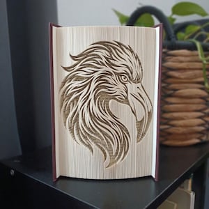 May include: A folded book art sculpture featuring the intricate profile of an eagle head. The pages are precisely creased and folded to create a three-dimensional relief of the bird's feathers, eye, and curved beak against the cream-coloured paper.