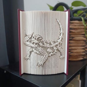 Könnte beinhalten: Eine Buchfaltkunst-Skulptur mit der detaillierten Silhouette einer Krötenechse, die durch präzises Falten der einzelnen Seiten eines Hardcover-Buches mit dunkelrotem Einband entstanden ist und die komplexe Textur der Schuppen zeigt.