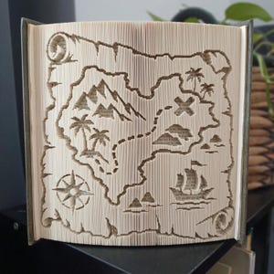 Può includere: Una scultura realizzata con le pagine di un libro piegate a formare una mappa del tesoro dettagliata. Il design include una rosa dei venti, palme, cime montuose, un veliero e una X che segna un punto, creati tramite precise tecniche di piegatura.