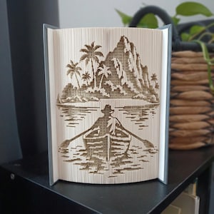 May include: A folded book art sculpture featuring a detailed paper-cut style design of a person rowing a small boat on water toward a tropical island with palm trees and a large mountain peak in the background, created by precisely folding book pages.