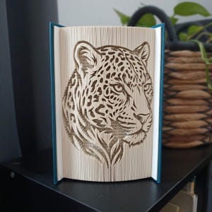 May include: A folded book art sculpture featuring a detailed leopard head design created by precisely folding the pages of a book. The sculpture displays the feline's spotted pattern and facial features through the layered edges of the paper.