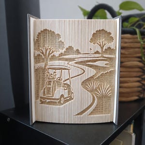 Peut inclure: Une sculpture en livre plié représentant une scène de terrain de golf. Les pages sont pliées avec précision pour révéler une voiturette de golf au premier plan, un chemin sinueux menant vers un green, des arbres et un drapeau au loin.