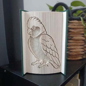 May include: A book sculpture featuring a folded page art design of a cockatoo parrot. The pages are precisely folded to create the silhouette of the bird, with the printed text of the book visible on the folded edges of the white paper pages.