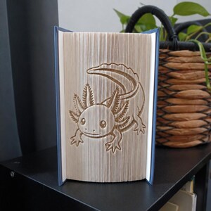 May include: A book art sculpture featuring a folded page design that creates a clear, three-dimensional image of an axolotl. The pages are fanned open to display the creature's outline, including its external gills, tail, and four limbs.