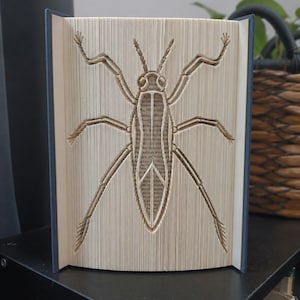May include: A book sculpture featuring folded pages that create a three-dimensional insect shape. The pages are cream-coloured with visible text, and the insect design is centred, showing a detailed body, head, antennae, and six long legs.
