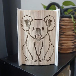 May include: A folded book art sculpture featuring a koala design created by precisely folding the pages of a book. The beige paper pages are manipulated to form the distinct outline of a koala with large ears, a round face, and a seated body.