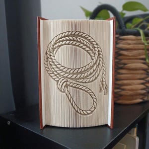 Puede incluir: Un libro con páginas dobladas que crean una escultura tridimensional de una cuerda enrollada con un lazo. El libro tiene una cubierta marrón y las páginas son de color crema, mostrando la intrincada técnica de plegado de papel.
