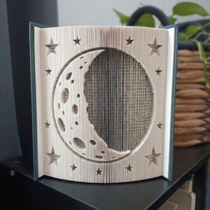 Könnte beinhalten: Eine Buchfaltkunst-Skulptur mit einem Mondsichel-Design inklusive Kratern und umgebenden Sternen. Die Seiten sind präzise gefaltet, um ein dreidimensionales Relief des Mondes zu erzeugen, wobei die dunkelgrünen Buchdeckel an den Seiten sichtbar sind.