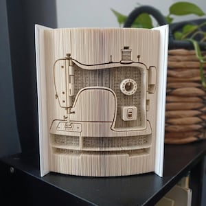 Puede incluir: Una escultura de libro con un diseño de páginas plegadas que crea la silueta de una máquina de coser. Las páginas son de color crema con texto impreso en negro visible, y los bordes plegados forman la forma detallada del cuerpo.
