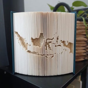 Könnte beinhalten: Eine Buchfaltkunst-Skulptur, die die Landkarte von Indonesien darstellt und durch präzises Falten der einzelnen Seiten eines Hardcover-Buches entsteht. Das Buch hat dunkelblaue Einbände und die weißen Papierkanten bilden die Silhouette.