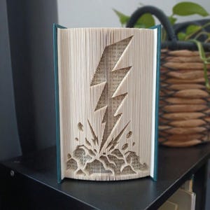 Peut inclure: Une sculpture en livre plié représentant un éclair, réalisée en pliant avec précision les pages d'un livre à couverture rigide. Les pages sont de couleur crème et le livre présente des couvertures bleu sarcelle foncé sur les bords.